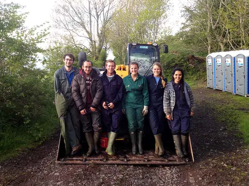 Six students in overalls and wellies standing in front of a yellow loader on a farm track