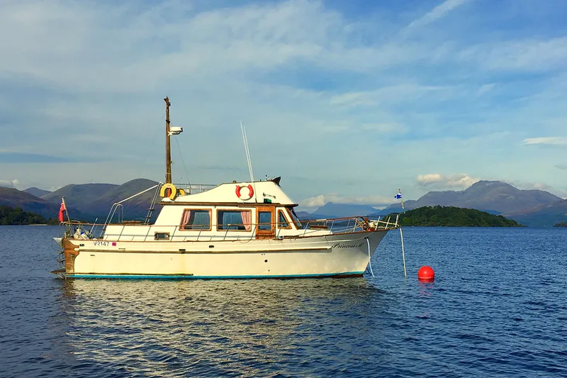 A white and cream cabin cruiser at anchor on blue water, with hills and a wooded island behind