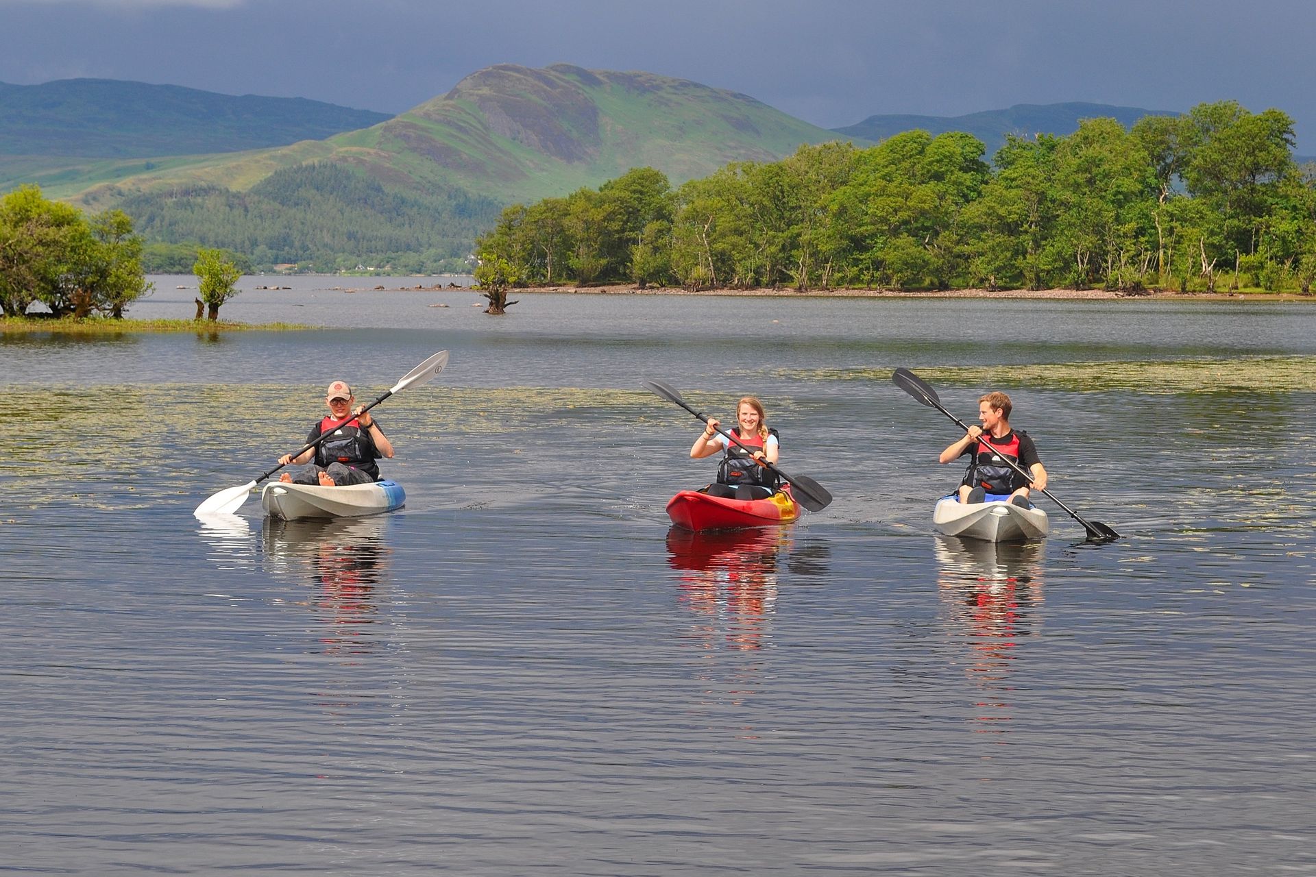 Three people paddling sit-on-top kayaks on calm water near a wooded shore, with hills behind