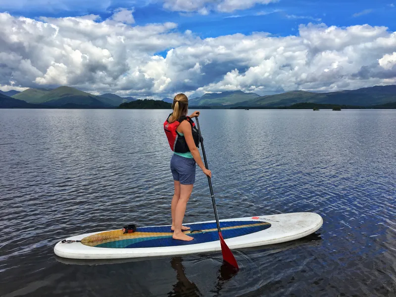 A woman stands on a stand-up paddleboard on Loch Lomond, with clouds and hills behind