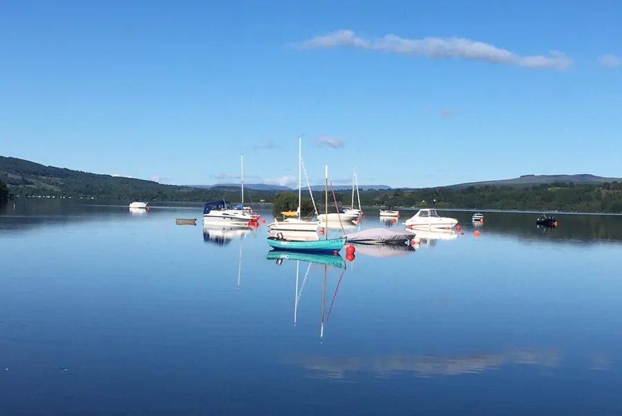 Moored boats on calm blue water in Portnellan Bay, with wooded hills across the loch and their reflections in the water
