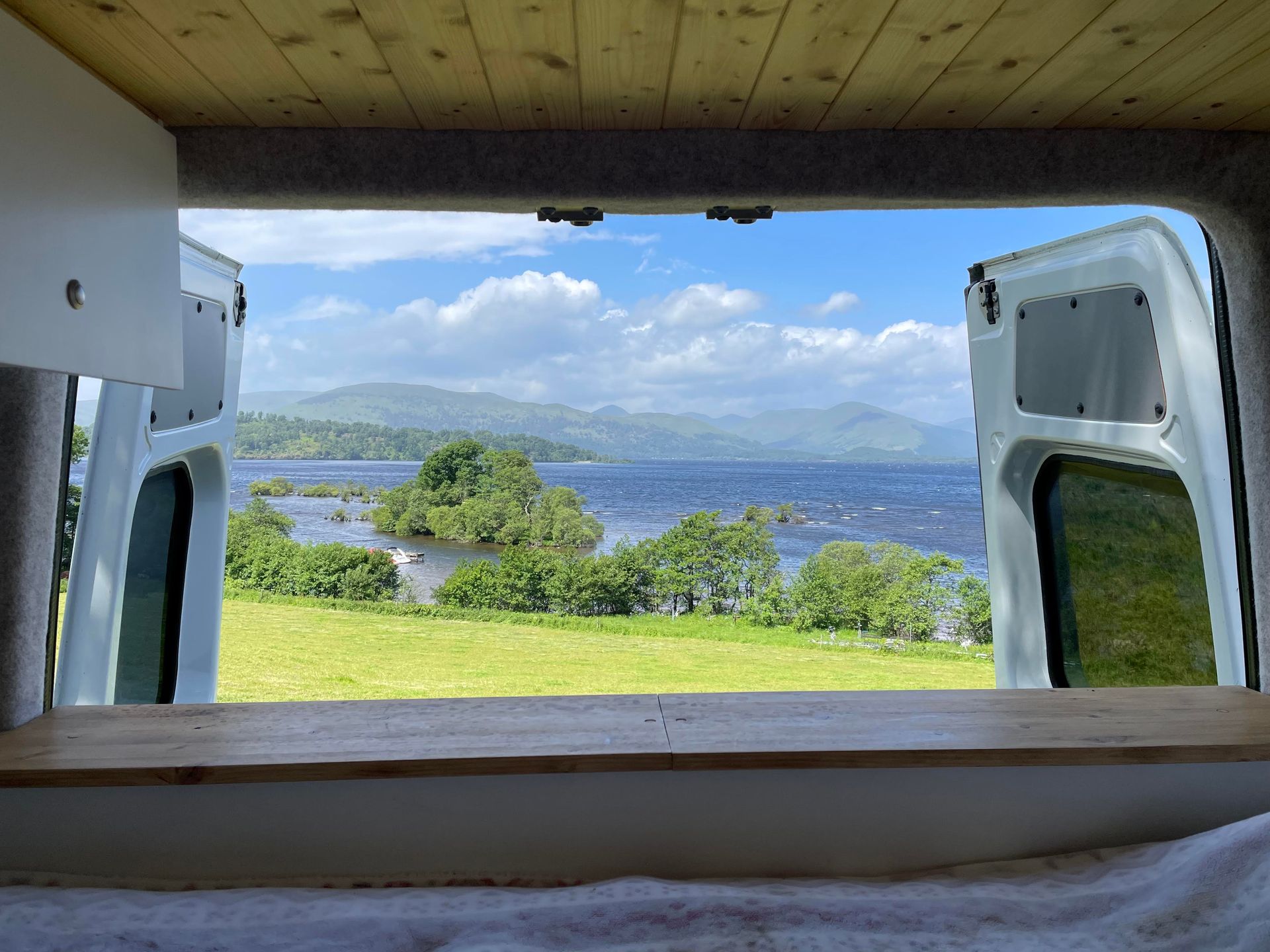 The open back doors of a campervan frame a view over fields and islands to Loch Lomond