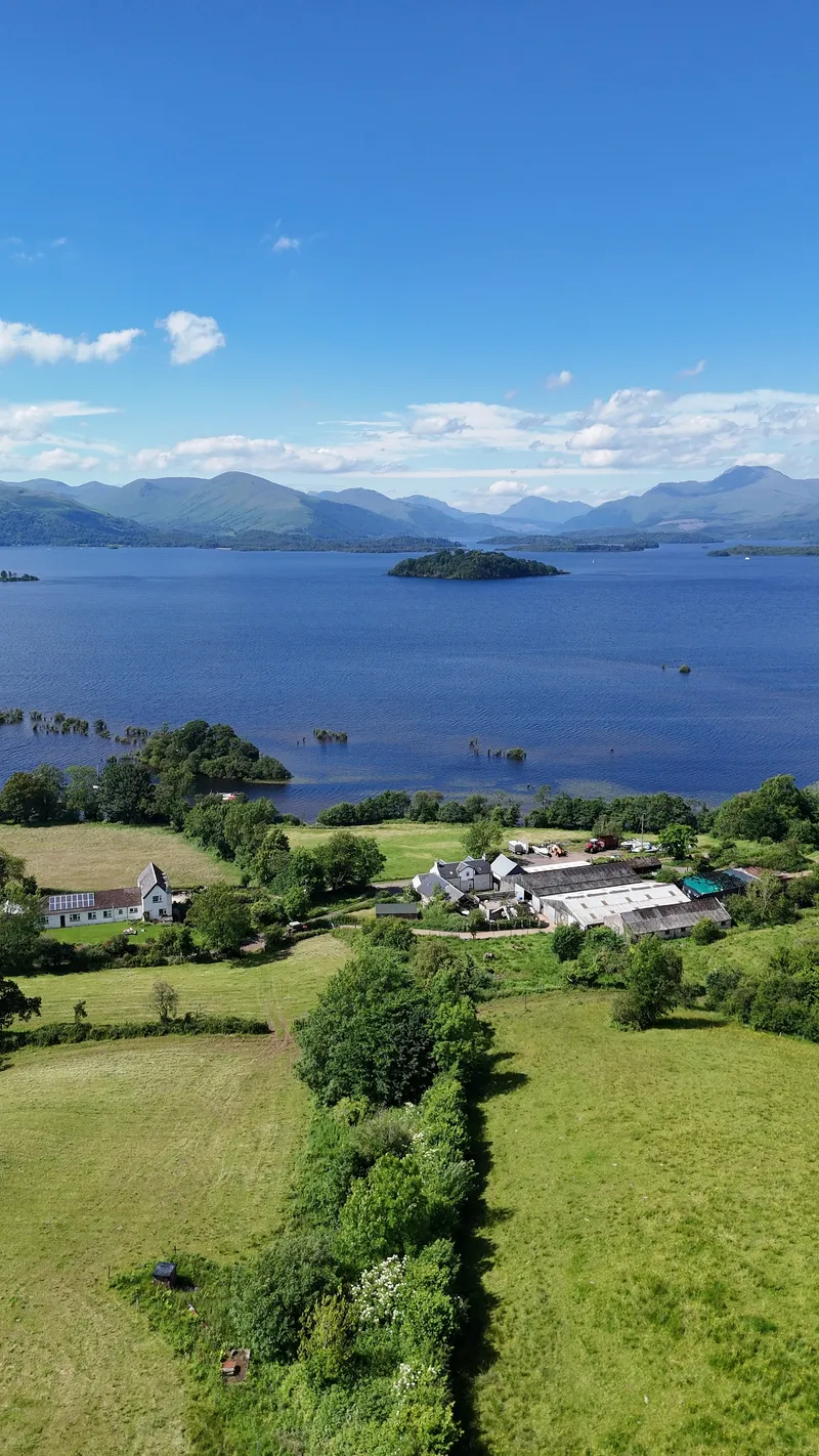 Aerial view over the fields and farm buildings at Portnellan towards Loch Lomond, with an island on the water and mountains beyond