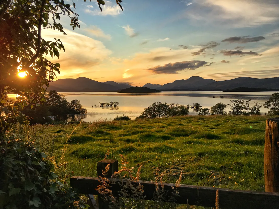 Sunset over Loch Lomond seen across a green field and a wooden fence, with islands on the water and hills beyond