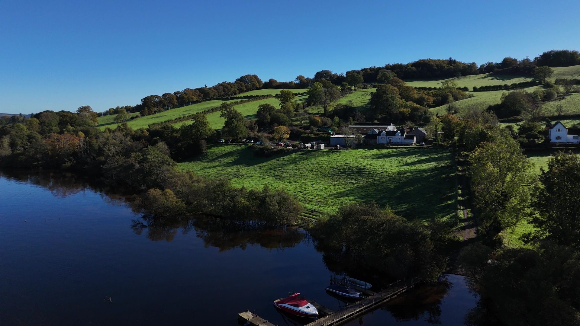 The farmhouse and farm buildings among green fields on a wooded hillside above the loch, with a boat at the jetty in the foreground