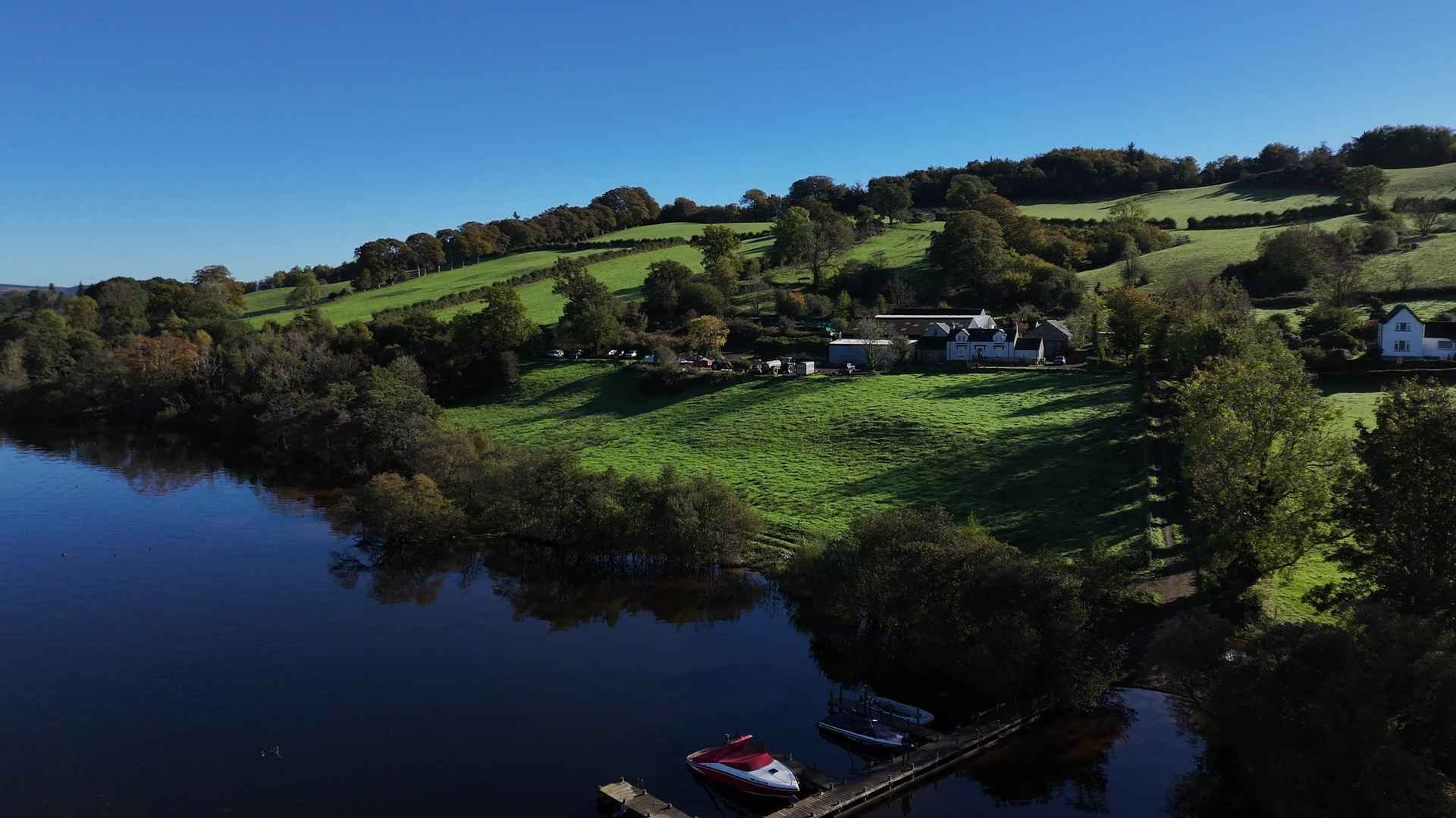 The farm's white buildings among green fields on a wooded hillside above the loch, with a boat moored at the jetty in the foreground