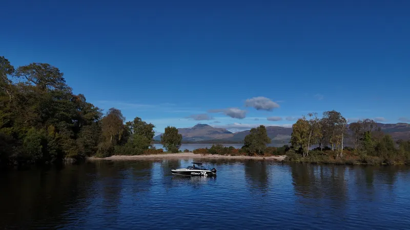 The Husky R8 on calm water beside a sandy beach on a wooded island, with HUSKY R8 on its hull and hills across the loch