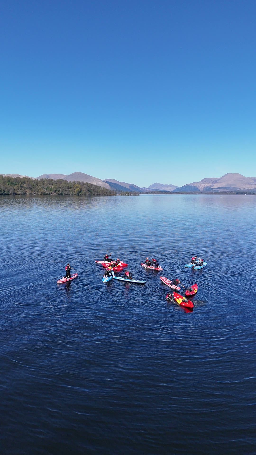 Kayakers and paddleboarders gathered on calm water on Loch Lomond, seen from above, with wooded hills beyond