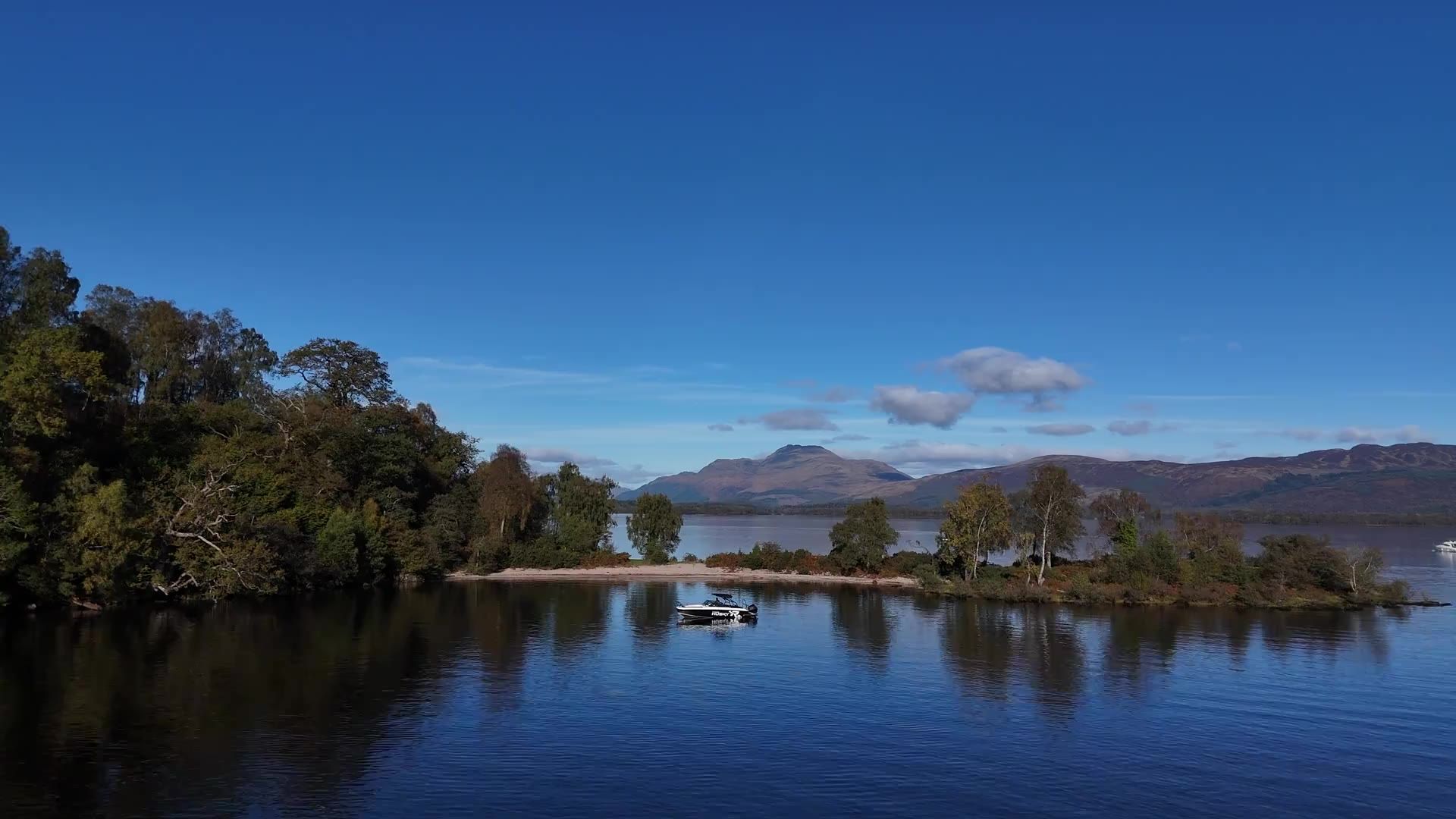 A speedboat on calm water off a sandy island beach, with trees on the shore and hills across Loch Lomond