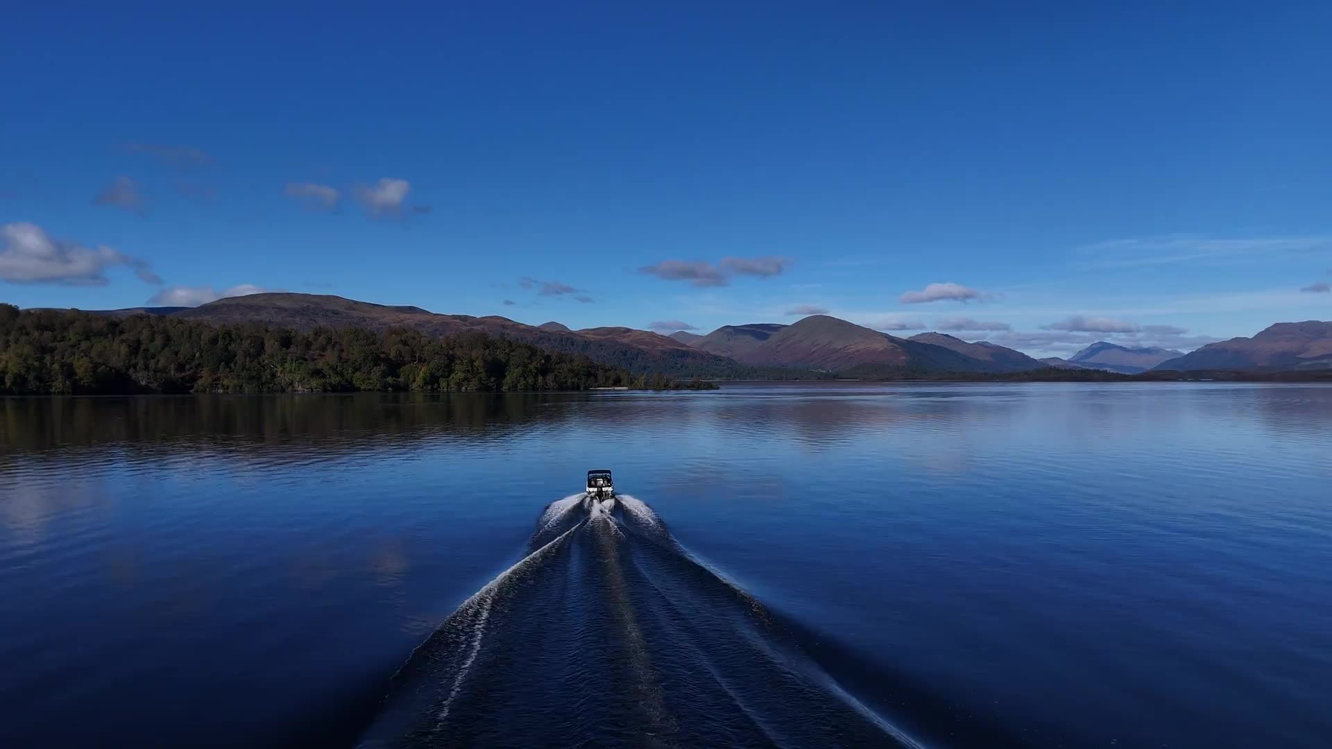 A speedboat seen from behind, leaving a long white wake across calm blue water towards the hills of Loch Lomond