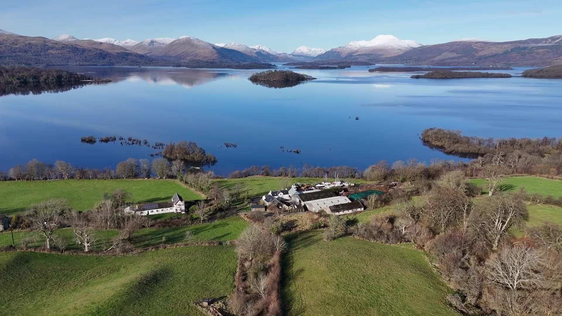 The farmhouse and farm buildings among green fields above Loch Lomond, with islands on the calm water and snow on the hills beyond